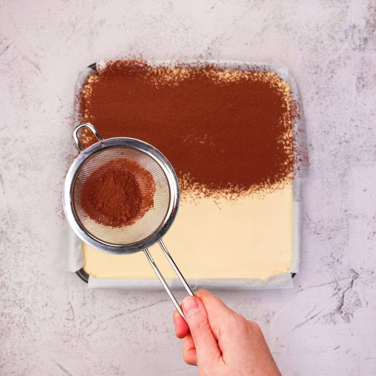 Fine cocoa powder being sifted over a vanilla and cocoa layer for an Italian dessert recipe.