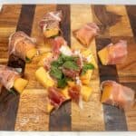 Thinly sliced prosciutto-wrapped cantaloupe and mozzarella cheese on a wooden serving board, perfect for Italian appetizers.