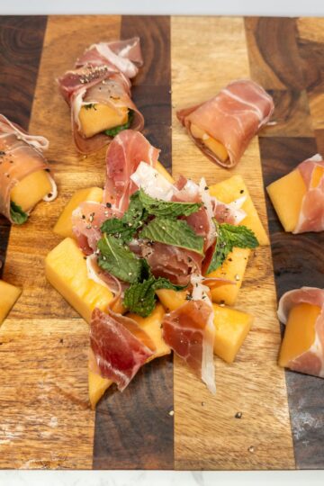 Thinly sliced prosciutto-wrapped cantaloupe and mozzarella cheese on a wooden serving board, perfect for Italian appetizers.