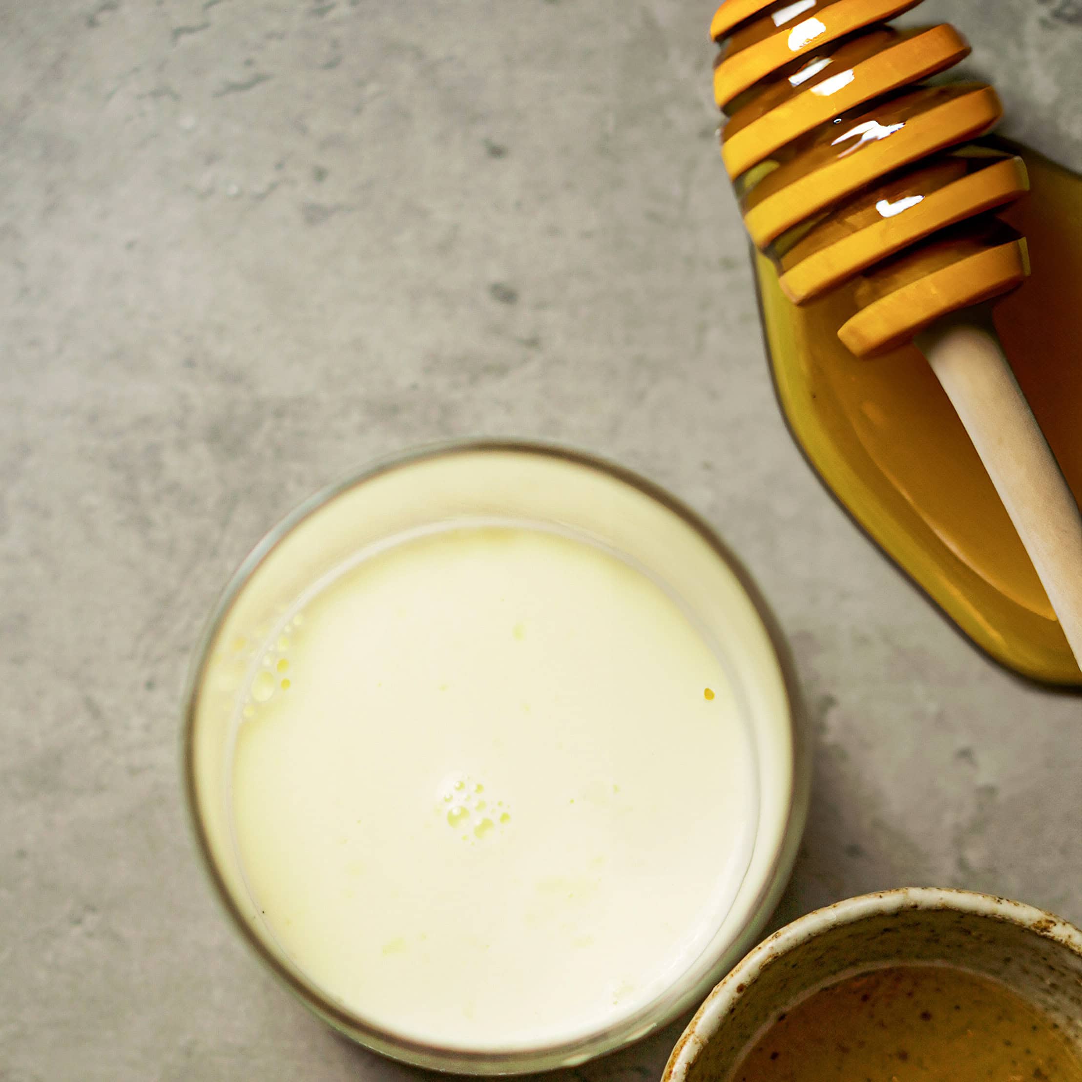 Glass of creamy honey and milk mixture with honey dipper on a neutral background.
